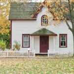 A charming white house with a red door and green shingles, surrounded by a white picket fence and autumn trees.