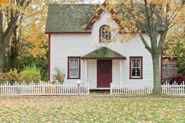 A charming white house with a red door and green shingles, surrounded by a white picket fence and autumn trees.